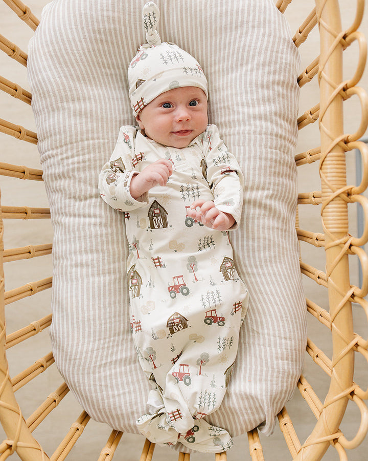 A baby wearing a cream-colored footed sleeper and matching hat with a tractor and barn pattern, lying on a striped cushion inside a wicker chair.