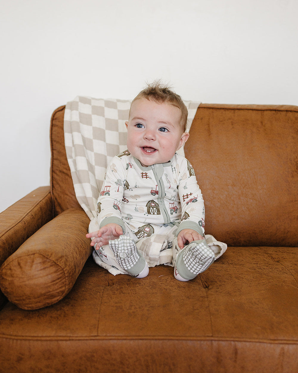 A baby sitting on a brown leather couch wearing a cream-colored zip-up sleeper with a brown and gray animal print and matching booties.