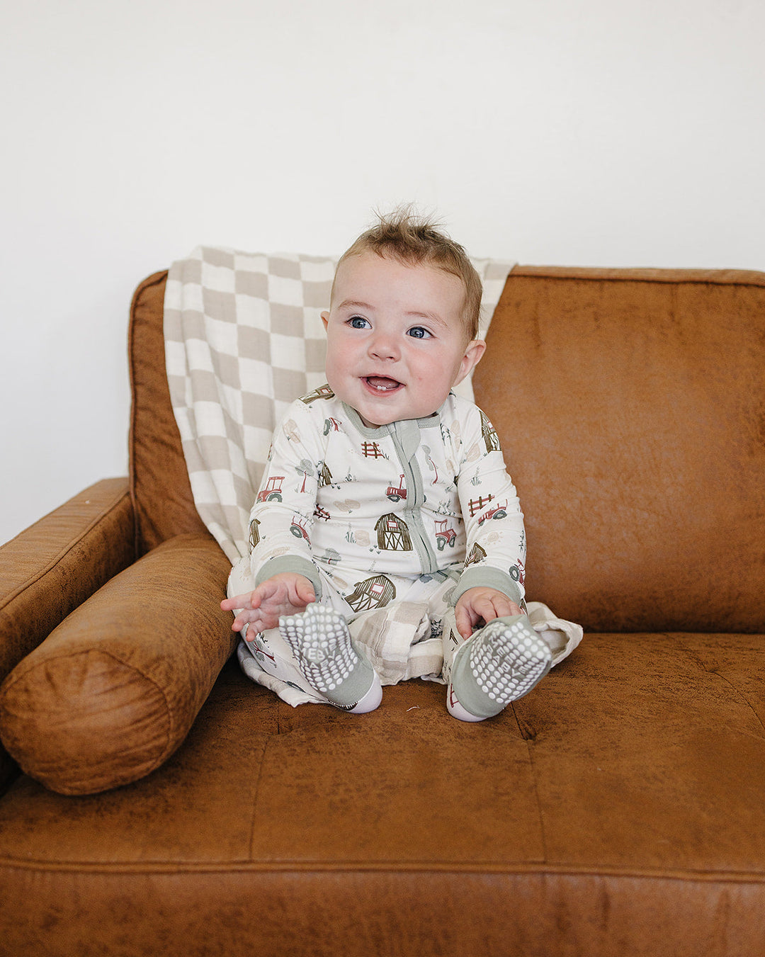 A baby sitting on a brown leather couch wearing a cream-colored zip-up sleeper with a brown and gray animal print and matching booties.
