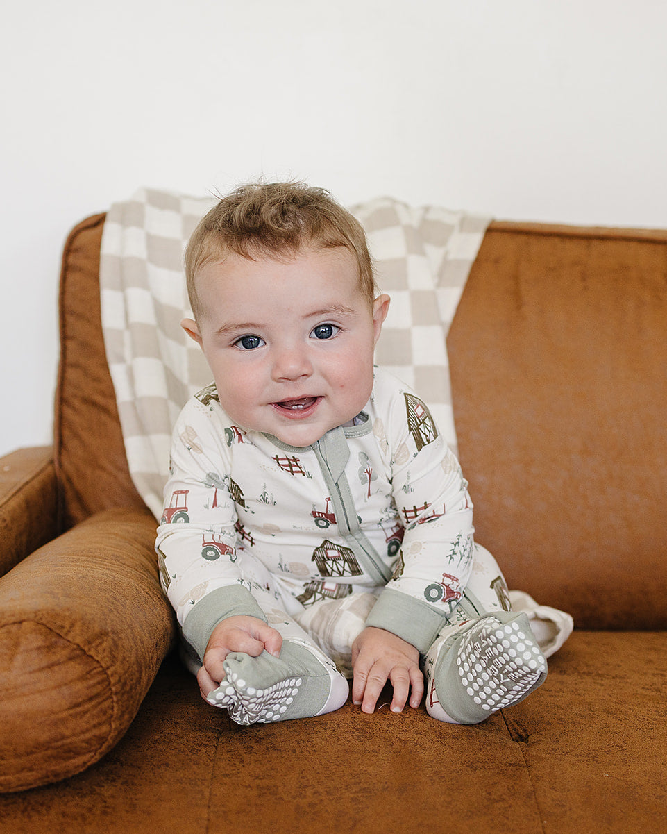 A baby sitting on a brown leather couch wearing a cream-colored onesie with a small truck pattern and gray booties.