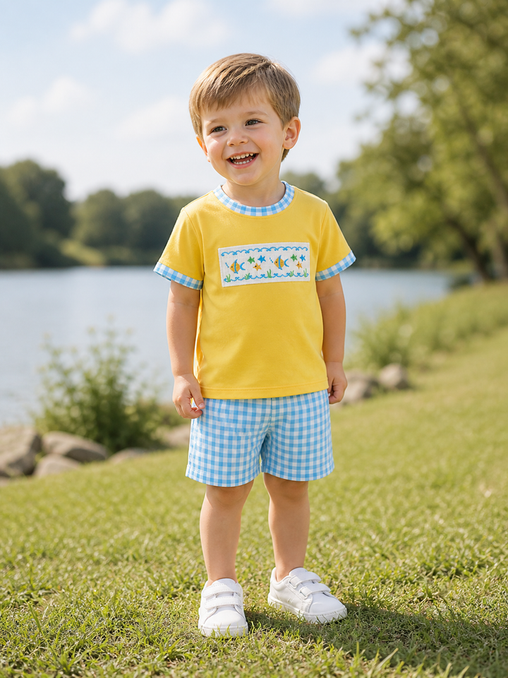 Child wearing a yellow shirt with a blue checkered pattern and white shoes, standing on grass by a lake.