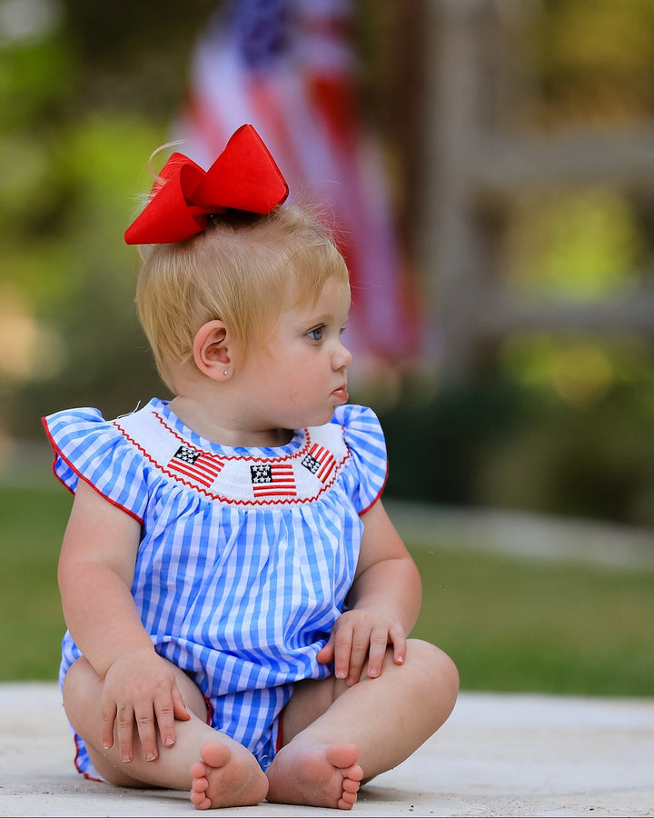 Baby in a blue and white checkered outfit with a red bow, sitting outdoors.