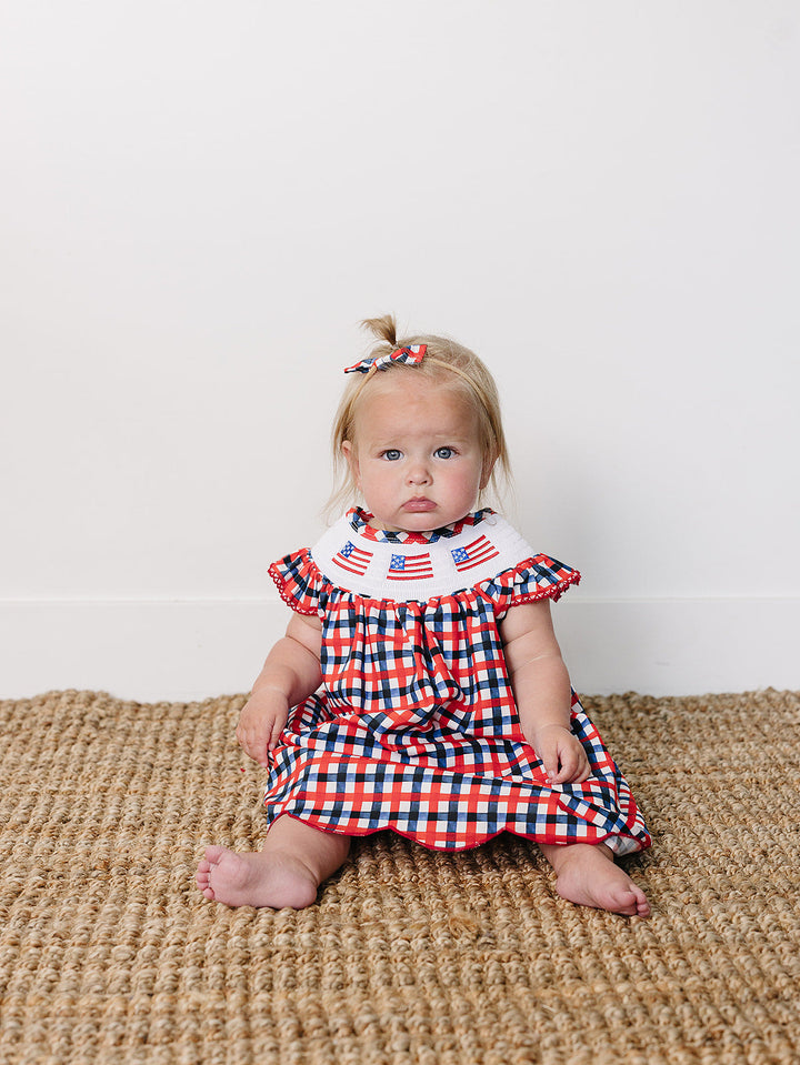 Baby wearing a red, white, and blue checkered dress with ruffled collar on a textured surface.