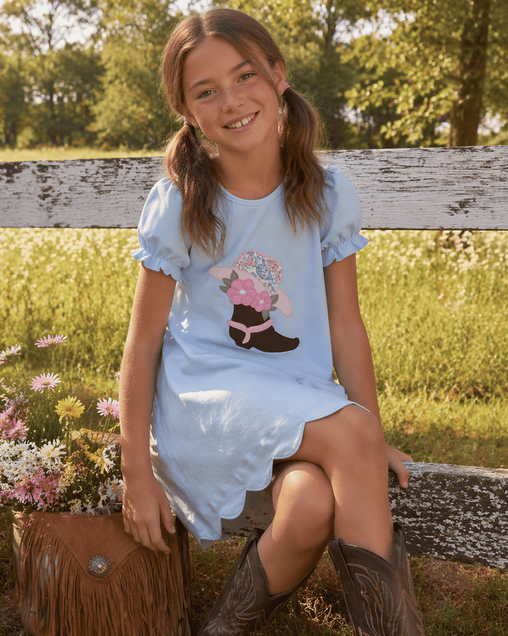 Young girl in a light blue dress with a floral boot design, sitting outdoors near a wooden fence and flowers.