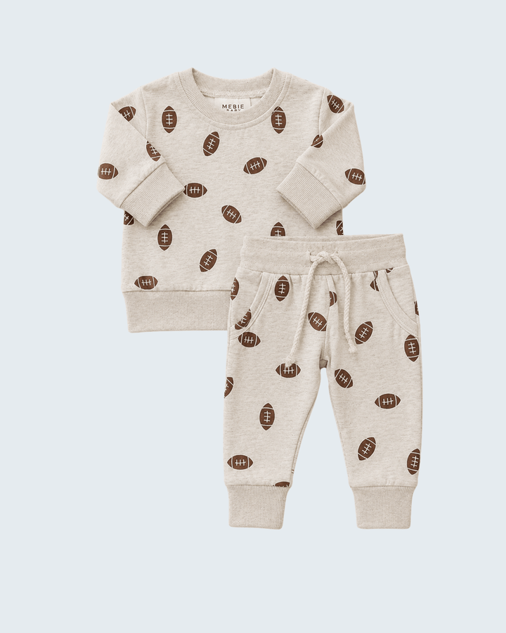 A baby two-piece loungewear set featuring a long-sleeve sweatshirt and matching jogger pants in a heather cream color, both printed with a brown football pattern, shown on a light blue-gray background.