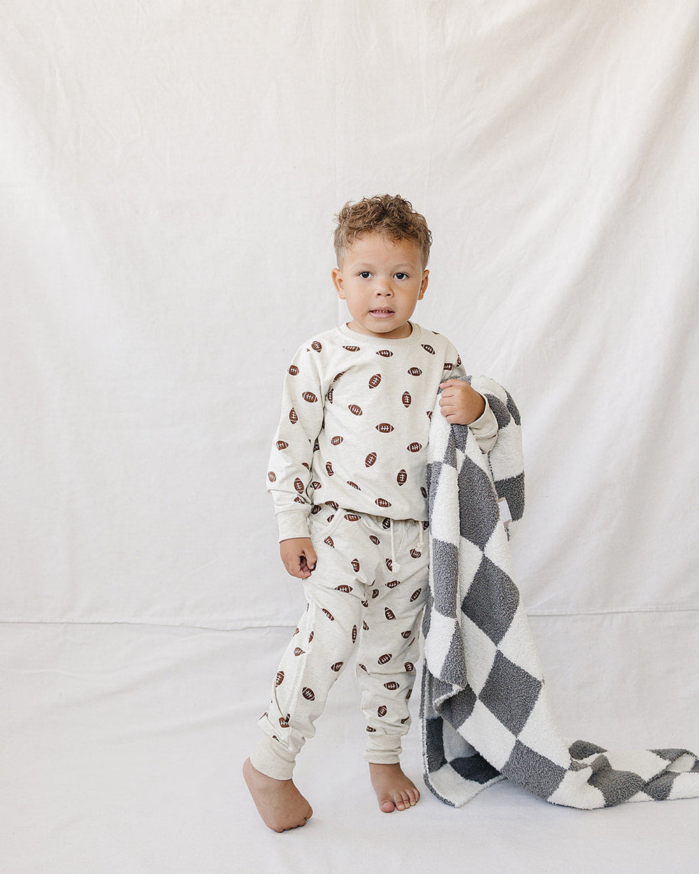 A toddler stands barefoot against a white fabric backdrop, wearing a cream long-sleeve pajama set with a brown coffee bean print and holding a black-and-white checkered blanket.