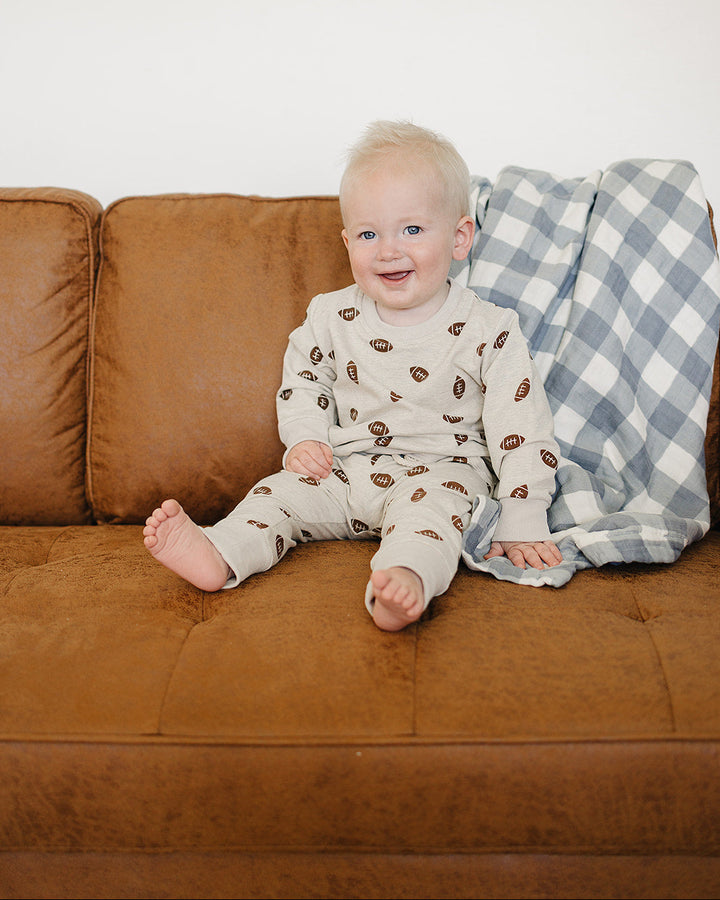 A baby sitting on a brown leather couch wearing a cream-colored one-piece jumpsuit with a brown leaf pattern, with a blue and white gingham blanket behind them.