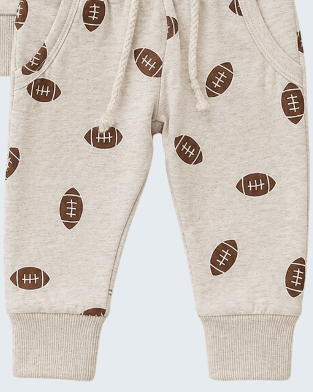 Close-up of a pair of baby jogger pants in a light heather cream color, featuring an all-over print of brown footballs with white laces and ribbed cuffs.
