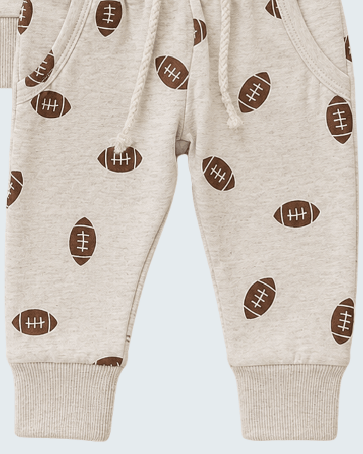 Close-up of a pair of baby jogger pants in a light heather cream color, featuring an all-over print of brown footballs with white laces and ribbed cuffs.
