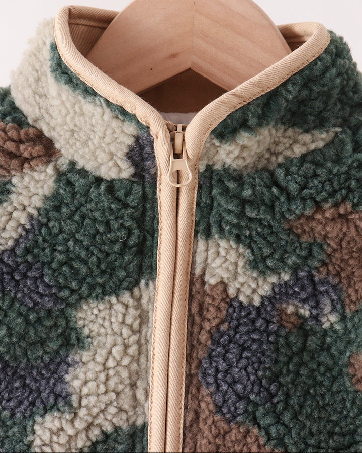 Close-up of a sherpa fleece zip-front jacket hanging on a wooden hanger, featuring a green, beige, cream, and brown camouflage pattern with beige trim.