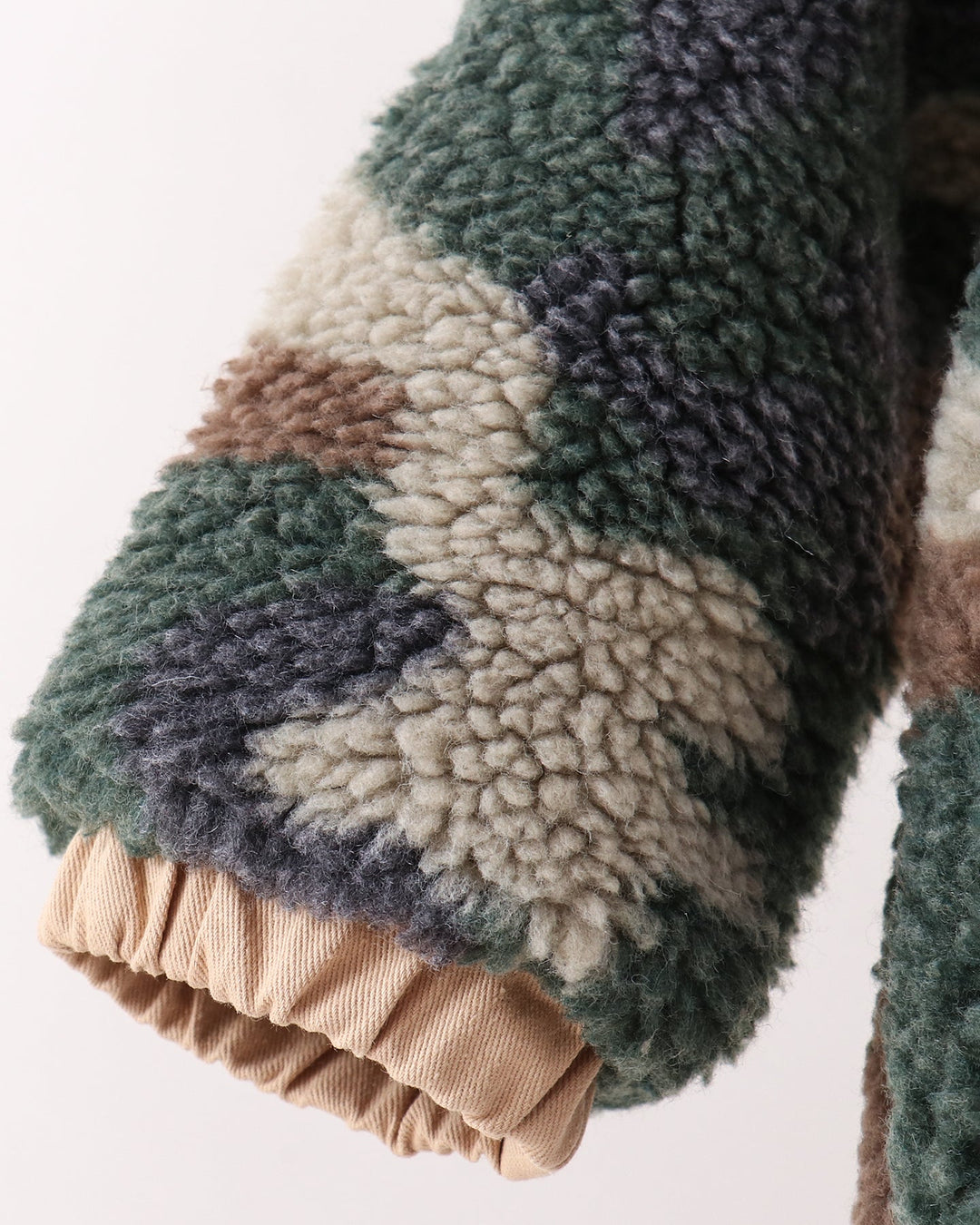 Close-up of a textured sherpa fleece garment sleeve with a camouflage pattern in green, beige, brown, and gray, finished with a gathered beige cuff.
