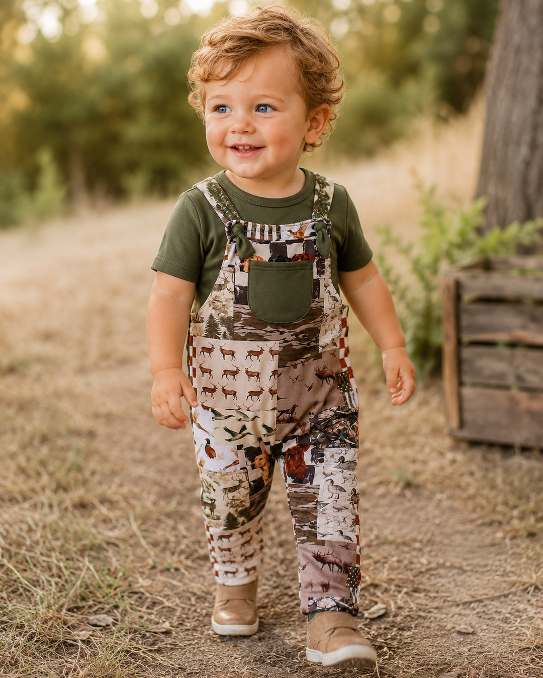 A toddler walking outdoors on a dirt path wearing a green short-sleeve t-shirt and brown camouflage patchwork overalls with dog-print bib panels, paired with tan shoes.