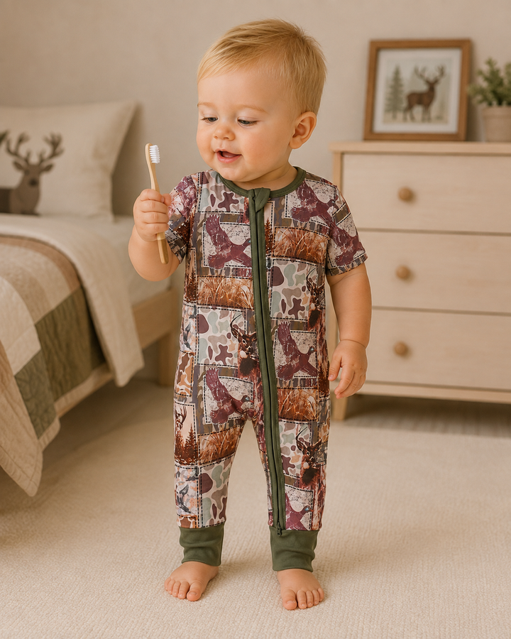 A baby standing on a carpeted floor in a nursery, wearing a short-sleeve zip-front footless onesie with a woodland animal print. The baby is holding a small toothbrush.