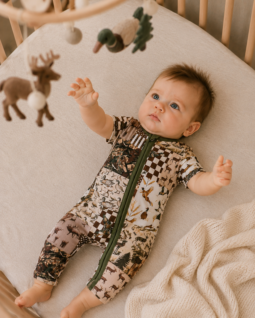A baby lying on their back in a crib, wearing a short-sleeve zip-front onesie with a woodland animal print. The print features brown, beige, and black tones with a dark green zipper.