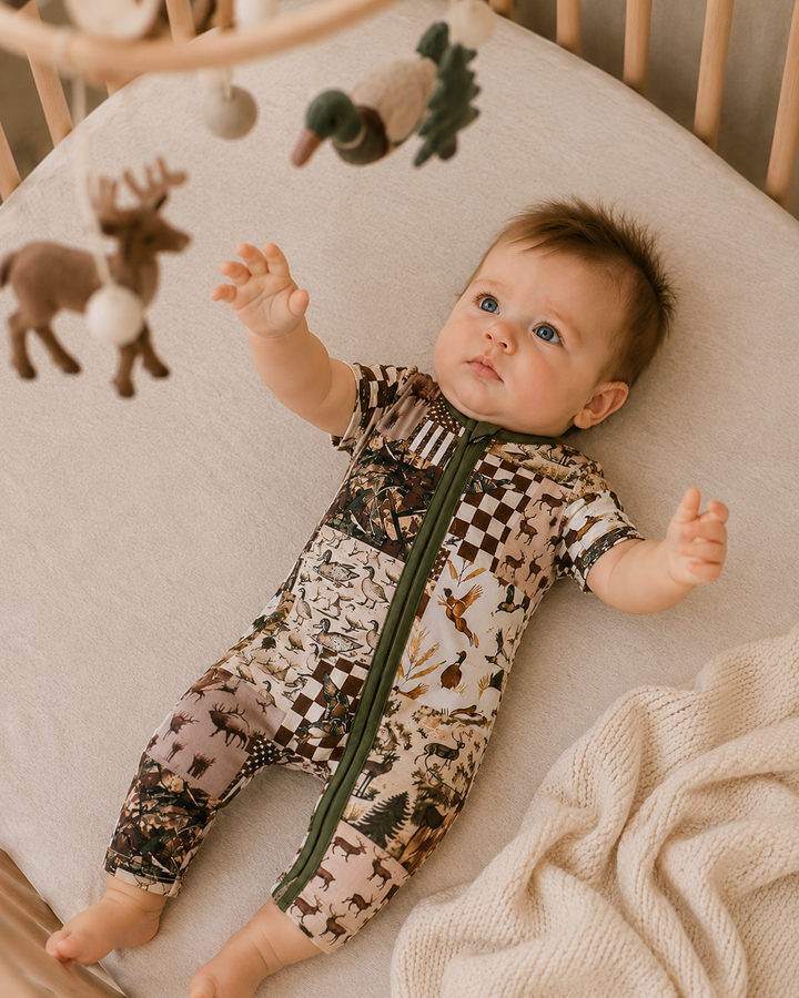 A baby lying on their back in a crib, wearing a short-sleeve zip-front onesie with a woodland animal print. The print features brown, beige, and black tones with a dark green zipper.