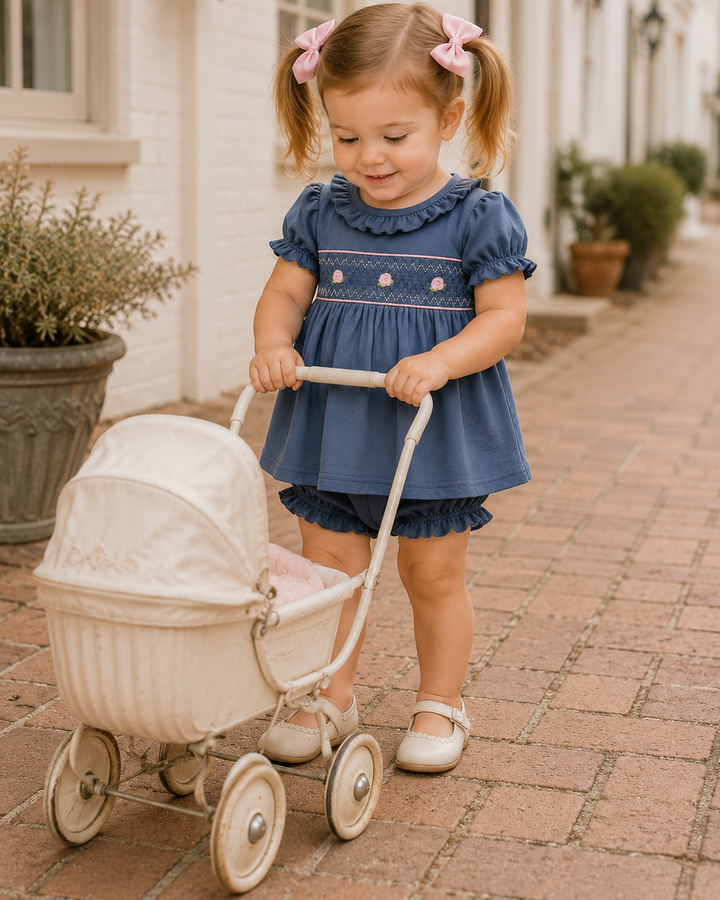 A young toddler girl with blonde pigtails wearing a blue smocked dress and white shoes pushes a cream-colored vintage toy carriage down a brick sidewalk in front of a white building.