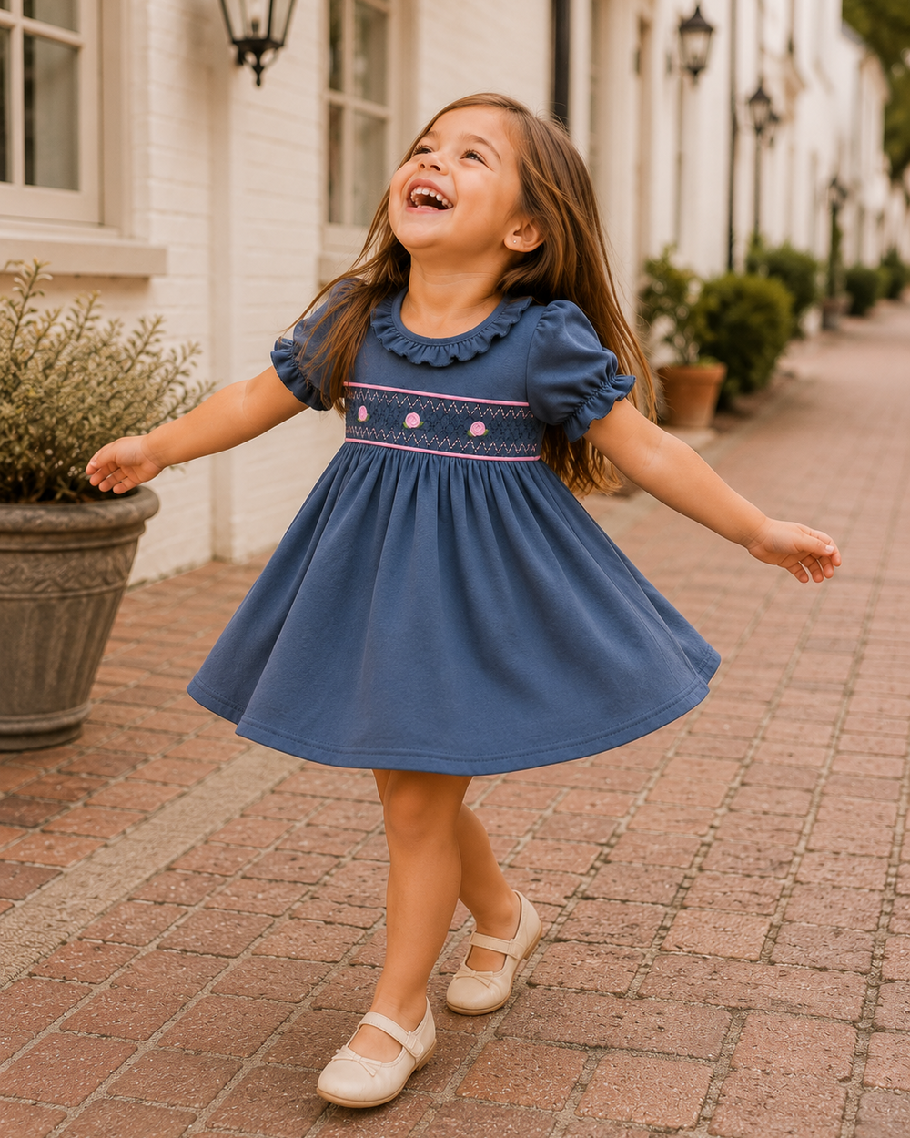 A young girl with long brown hair is captured mid-spin on a brick sidewalk, wearing a blue short-sleeve dress with pink and white embroidery across the chest and cream-colored shoes. The background shows a sunny, out-of-focus street scene with white buildings and potted plants.