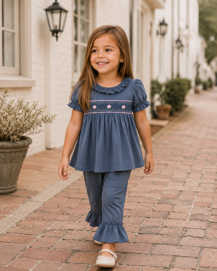 A young girl walking on a brick sidewalk in front of a white building, wearing a dusty blue two-piece outfit with a ruffled collar and smocked bodice, matching ruffled pants, and white shoes.