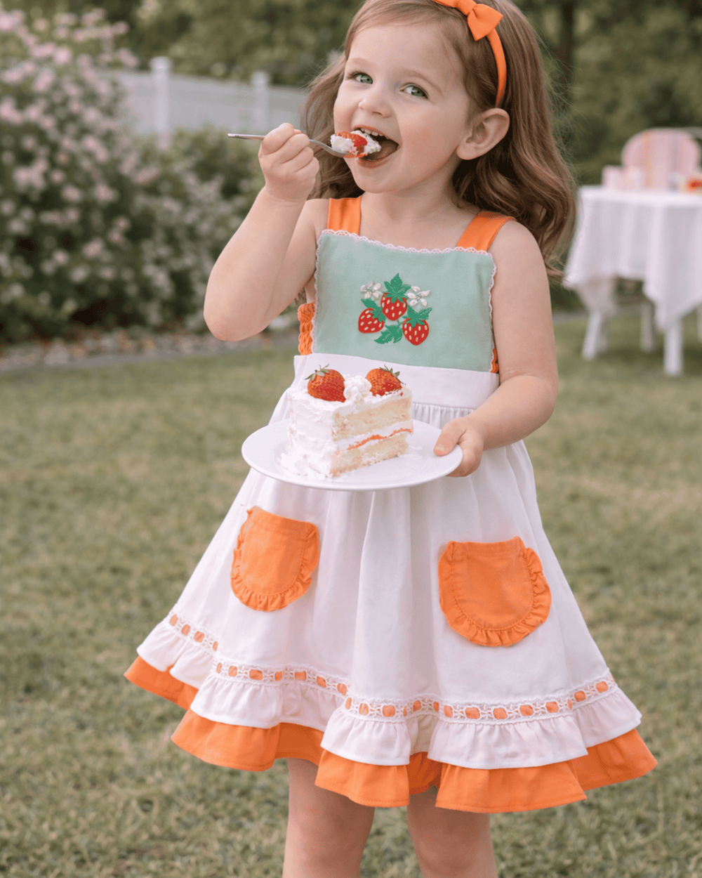 Young girl in a dress with strawberry design eating cake outdoors.