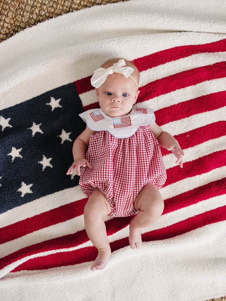 Baby in a red checkered outfit with a white bow on an American flag blanket