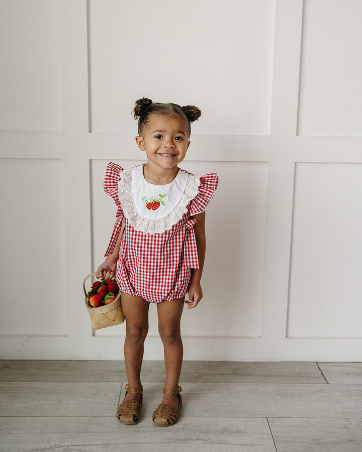 Young girl in a red checkered outfit holding a basket of fruits against a white paneled wall.