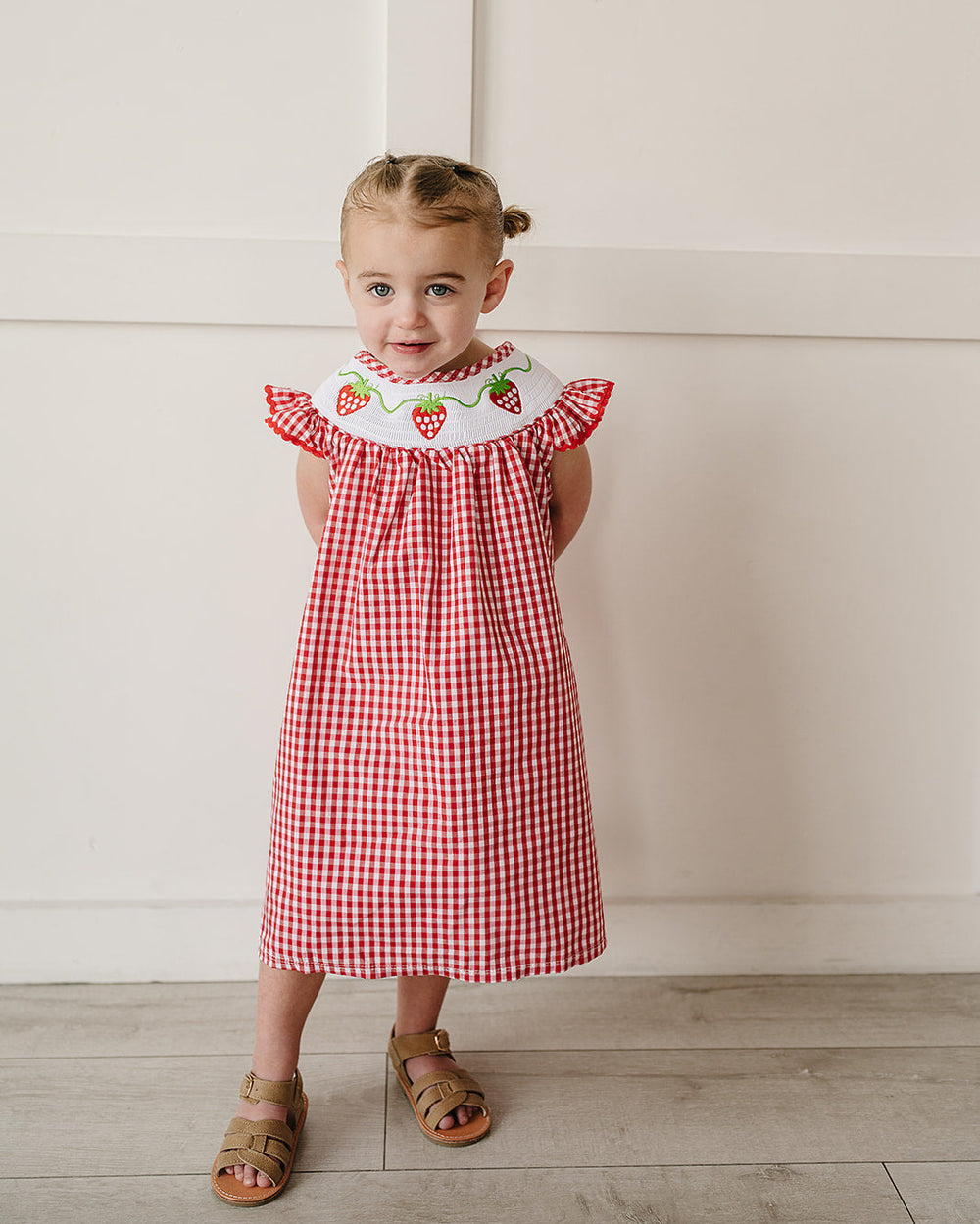 Young girl wearing a red checkered dress with strawberry details against a white wall.