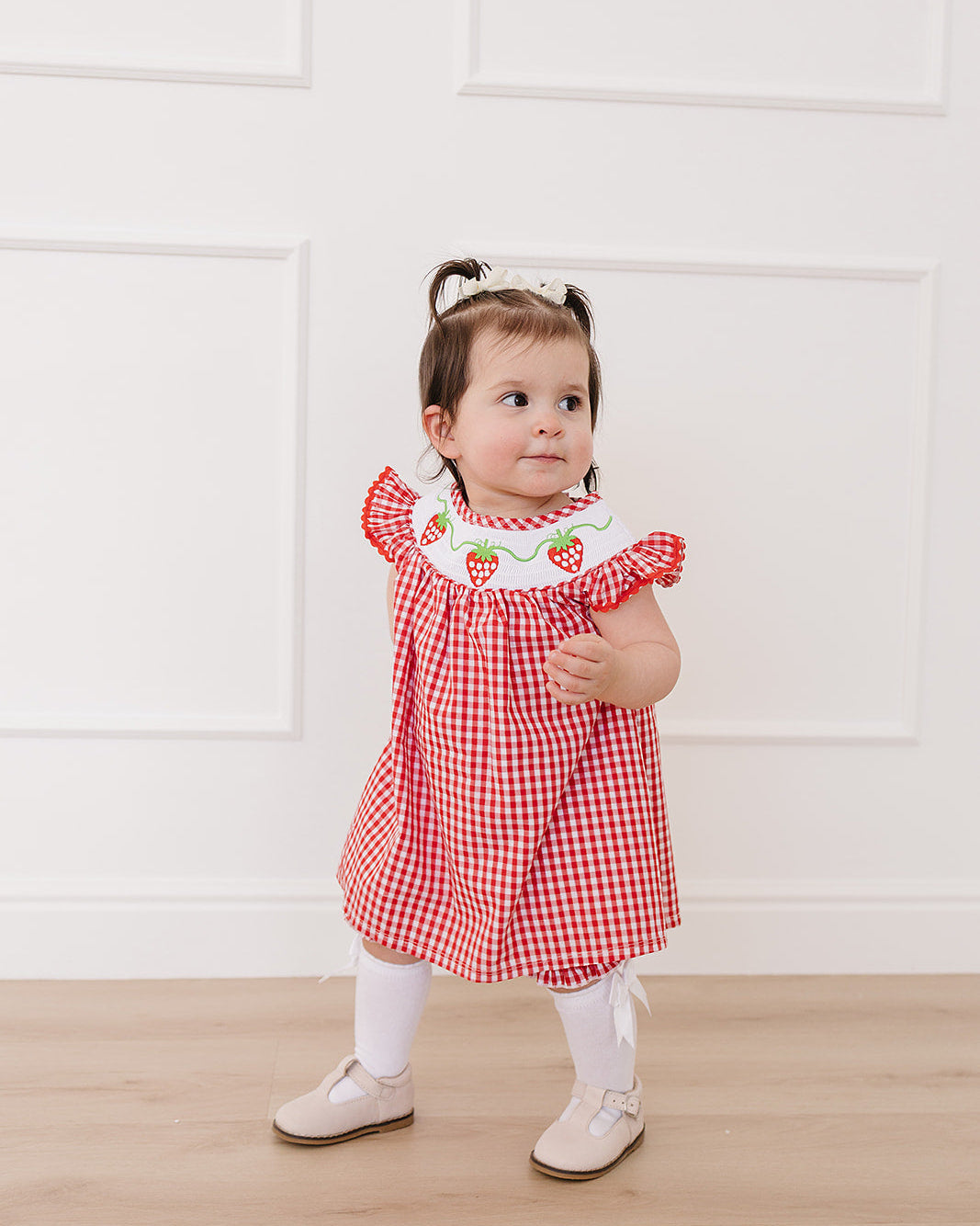 Child wearing a red checkered dress with strawberry details against a white wall.