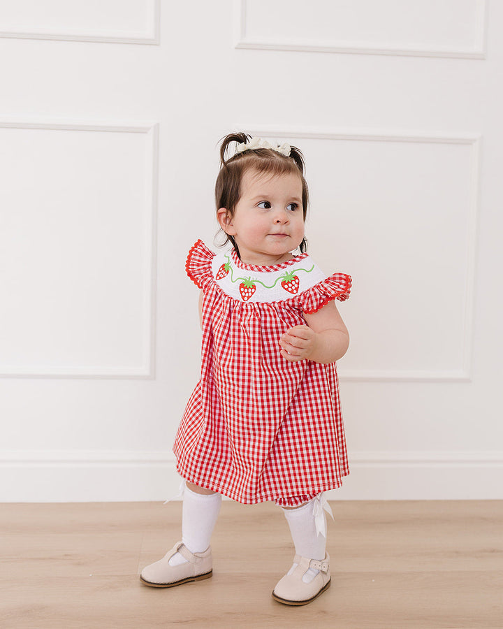 Child wearing a red checkered dress with strawberry details against a white wall.