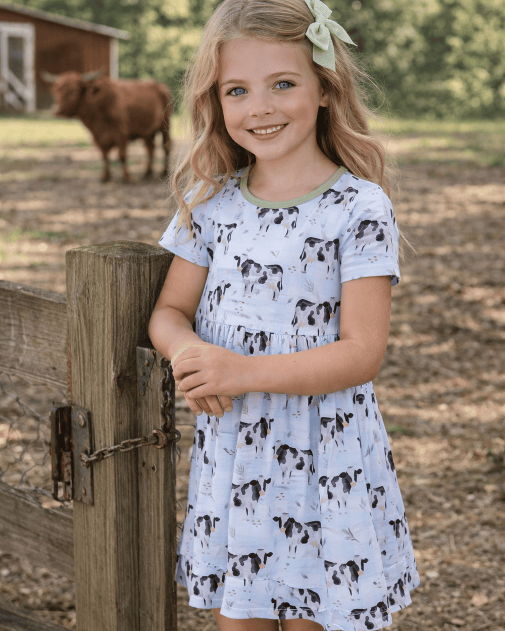 Young girl in a cow-print dress standing next to a wooden fence with a cow in the background.