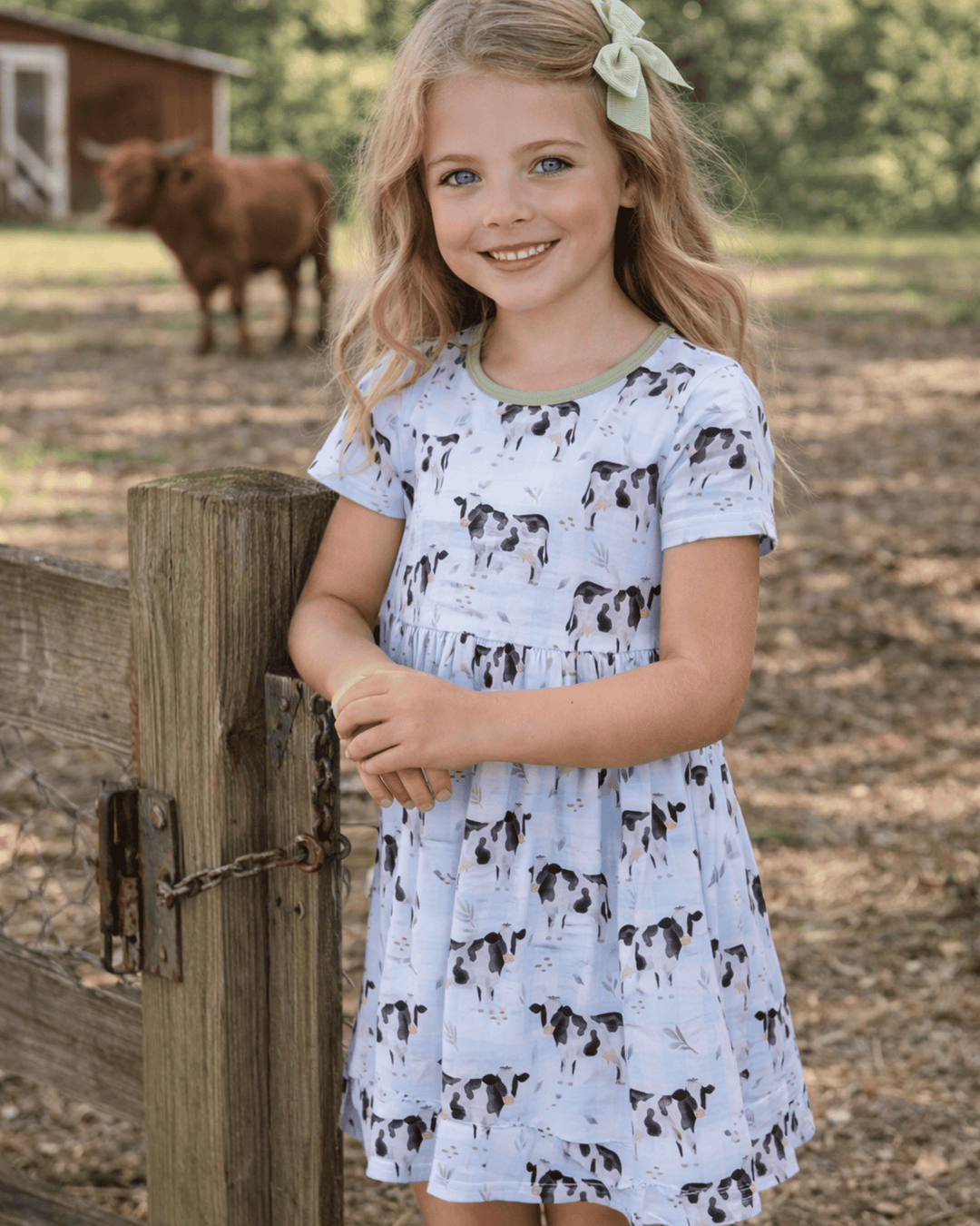 Young girl in a cow-print dress standing next to a wooden fence with a cow in the background.