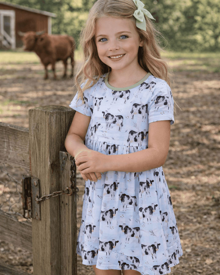Young girl in a cow-print dress standing next to a wooden fence with a cow in the background.