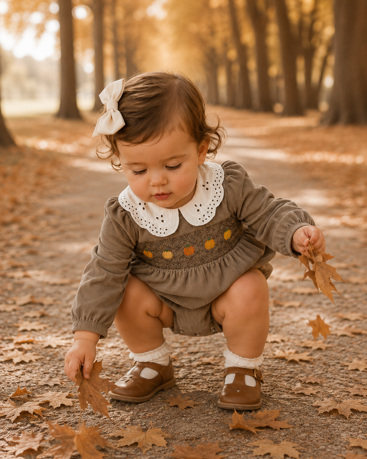 A toddler crouches on a leaf-strewn path in a wooded area, wearing a brown long-sleeve romper with a white Peter Pan collar, brown shoes, white socks, and a white bow in her hair.