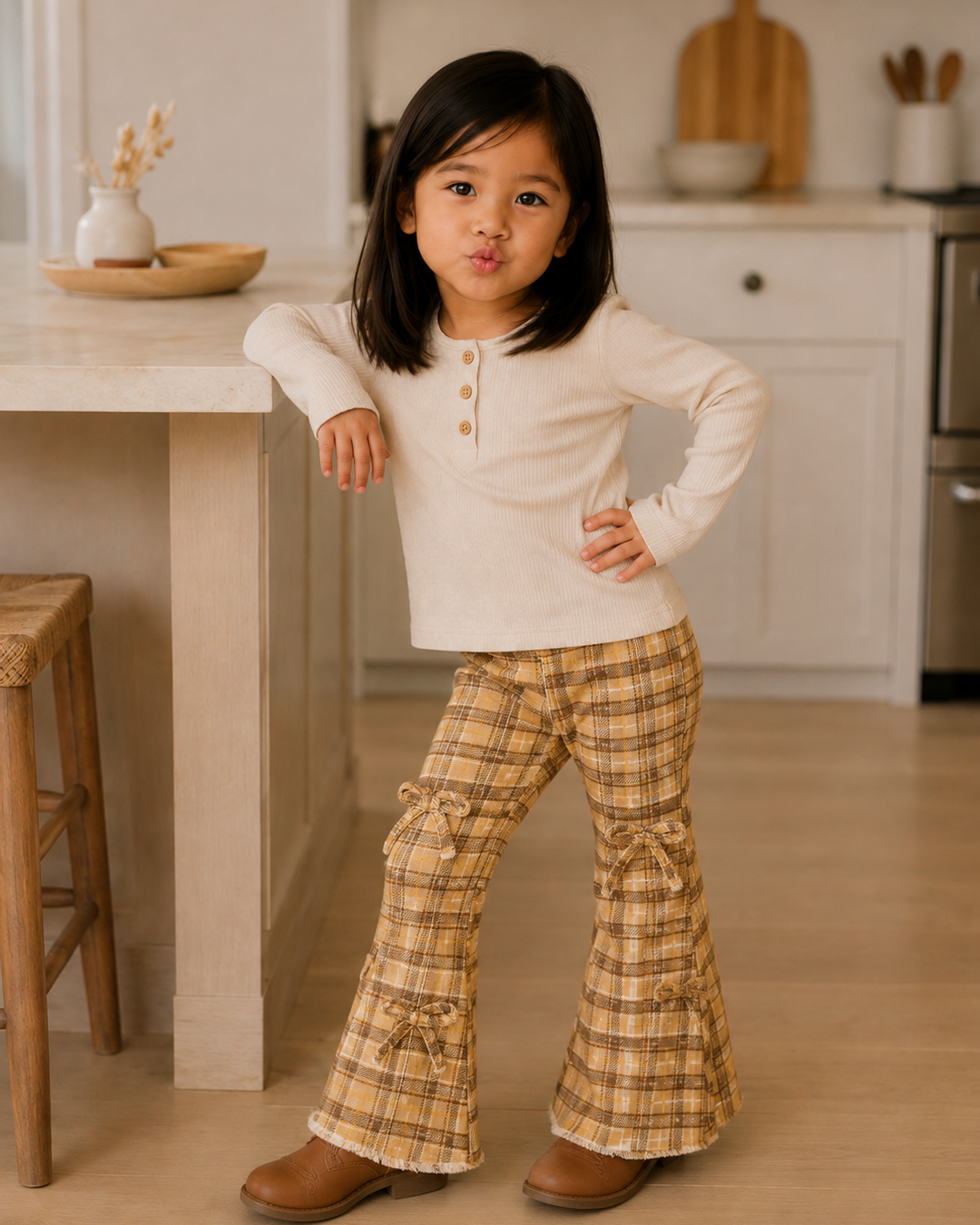 A young girl with shoulder-length dark hair stands in a bright kitchen, wearing a cream long-sleeve henley top and tan-and-cream plaid flared pants with bow details at the cuffs, paired with brown boots.