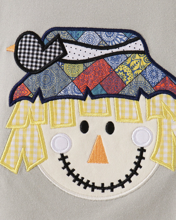 A close-up of a colorful embroidered scarecrow applique on a light gray fabric background. The applique features a patchwork patterned hat in blue, red, yellow, and black, yellow gingham hair, a cream-colored face, and a stitched smile.