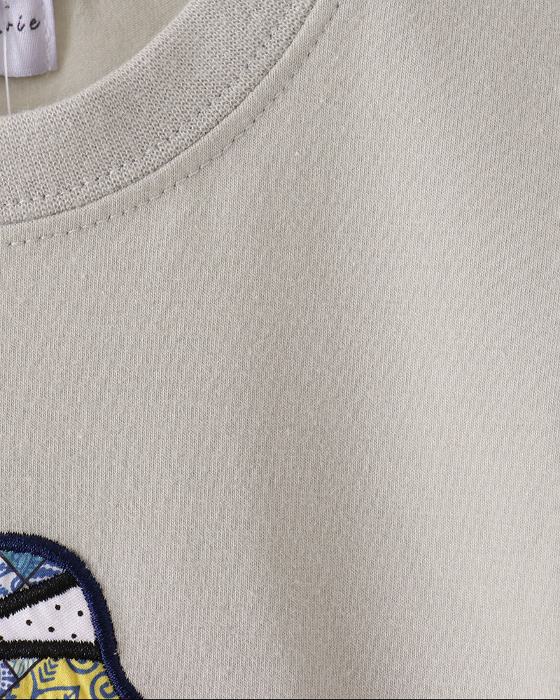 Close-up of a light beige crewneck sweatshirt on a wooden hanger, showing the ribbed collar and a partial embroidered patch with blue, yellow, and white details.