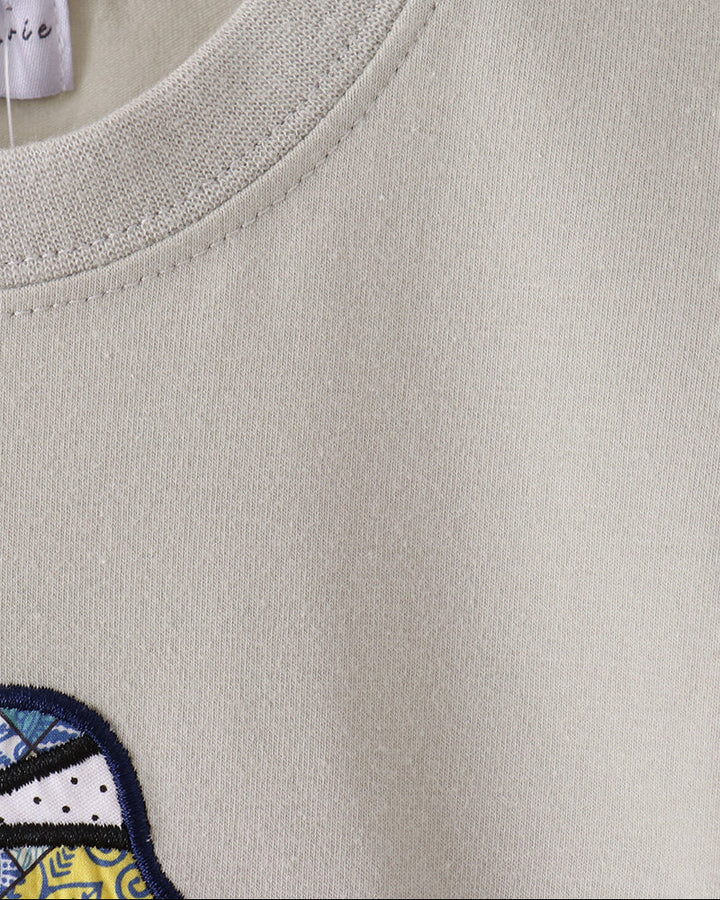Close-up of a light beige crewneck sweatshirt on a wooden hanger, showing the ribbed collar and a partial embroidered patch with blue, yellow, and white details.