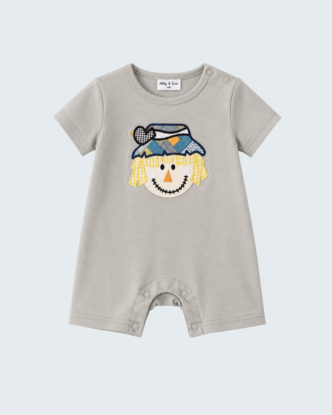 A baby short-sleeve romper in light beige, featuring a colorful embroidered scarecrow face on the chest. The romper has a crew neck, snap closures at the crotch, and a white brand label inside the collar that reads 'Milly & Tom'.