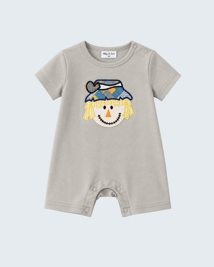 A baby short-sleeve romper in light beige, featuring a colorful embroidered scarecrow face on the chest. The romper has a crew neck, snap closures at the crotch, and a white brand label inside the collar that reads &
