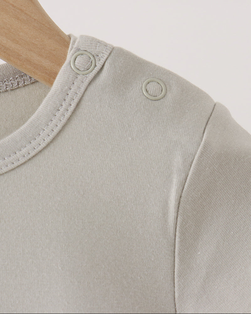 Close-up of a light beige baby top on a wooden hanger, showing the shoulder with two circular snap closures and fine ribbed knit fabric.