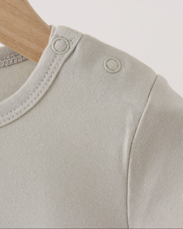 Close-up of a light beige baby top on a wooden hanger, showing the shoulder with two circular snap closures and fine ribbed knit fabric.