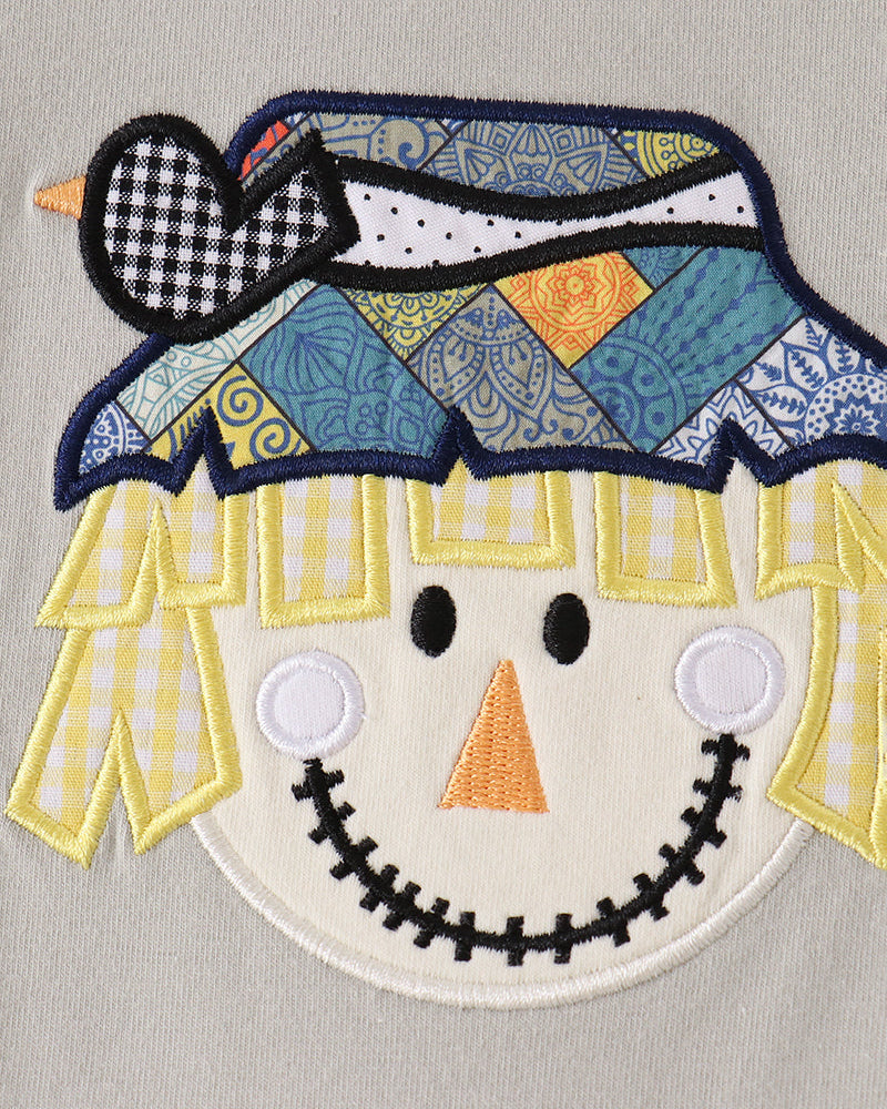 A close-up of a patchwork-style embroidered scarecrow face applique on a light gray fabric background. The hat is made of blue, yellow, and white patterned fabric sections with a black-and-white check heart accent.