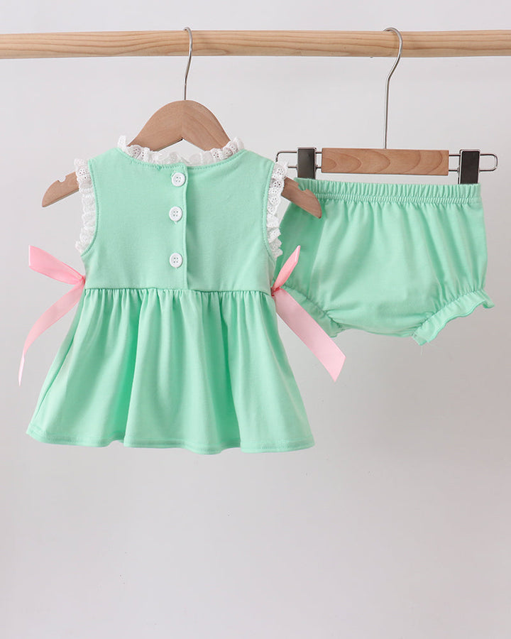 Mint green baby dress with buttons and pink ribbons on a wooden hanger against a light gray background.