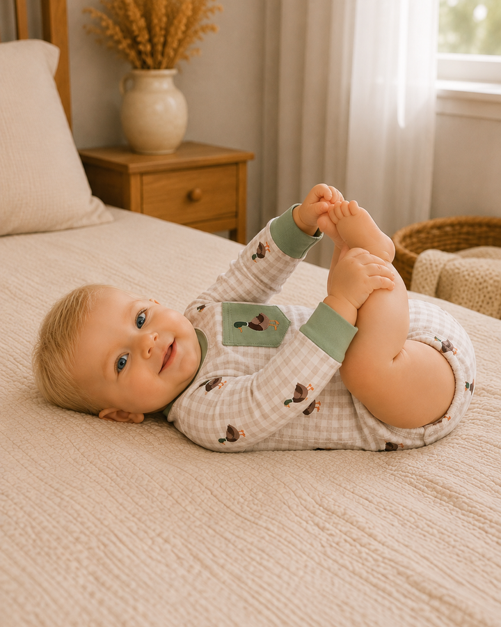 A baby lying on a textured cream bedspread, wearing a light beige and white gingham long-sleeve romper with sage green cuffs and a green chest patch. The background shows a cozy bedroom setting with a wooden nightstand, a vase of dried grass, and a window with sheer curtains.