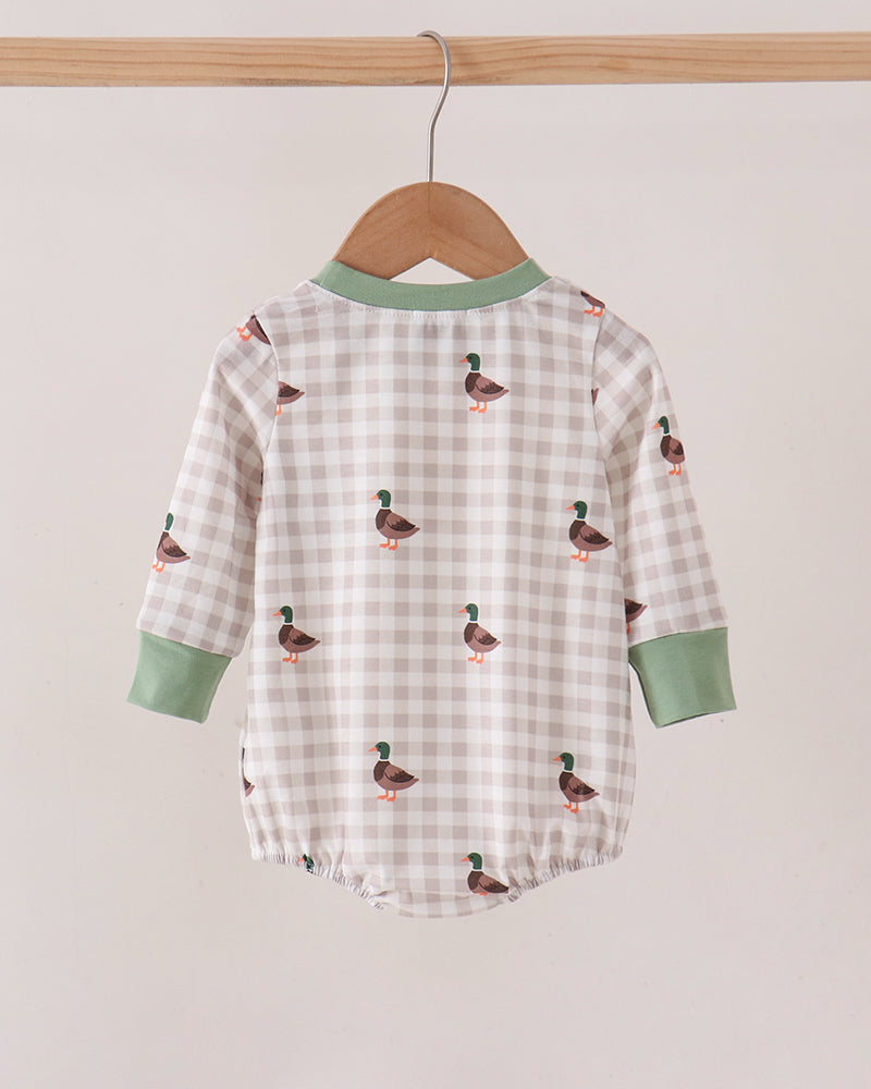 A baby long-sleeve romper hanging on a wooden hanger against a plain light beige wall. The romper has a white and beige gingham pattern with small duck prints, sage green collar and cuffs, and is shown from the back.