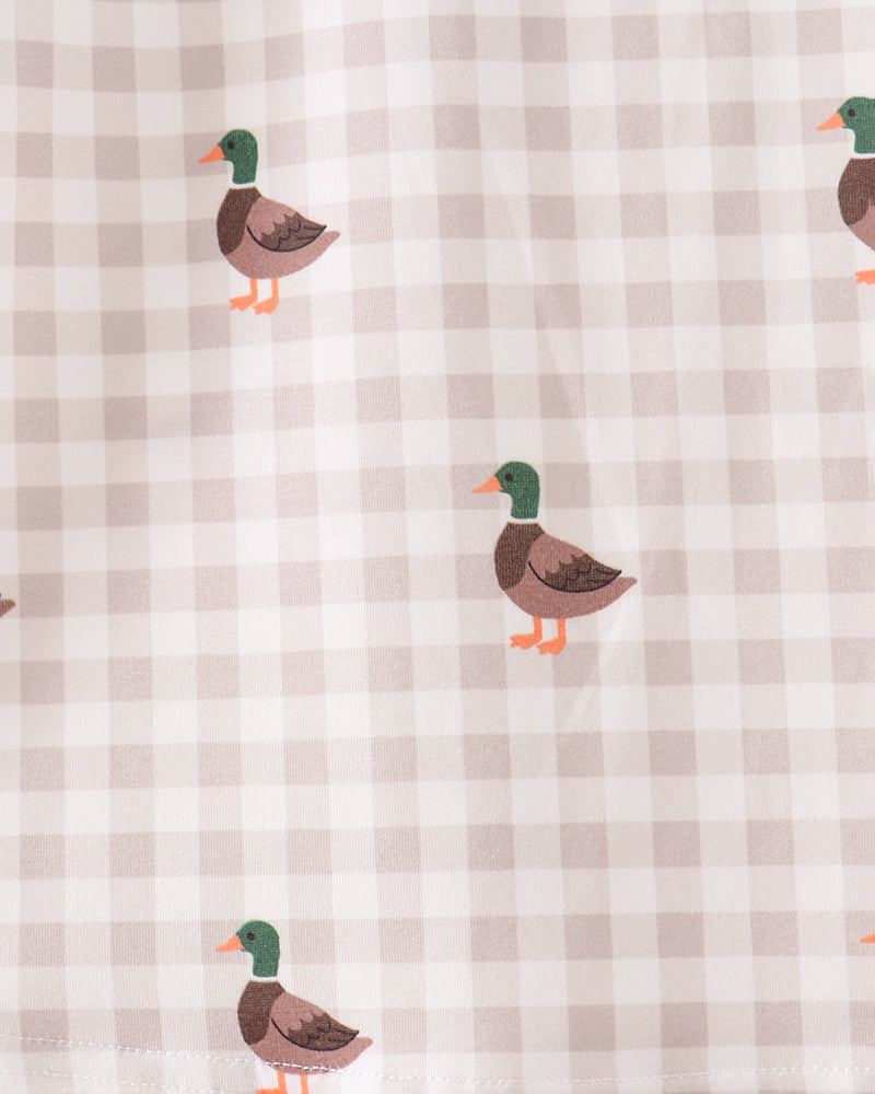 A close-up of a fabric surface with a light beige and white gingham check pattern and scattered duck illustrations. The ducks have green heads, brown bodies, and orange beaks and feet.