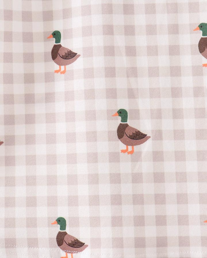 A close-up of a fabric surface with a light beige and white gingham check pattern and scattered duck illustrations. The ducks have green heads, brown bodies, and orange beaks and feet.