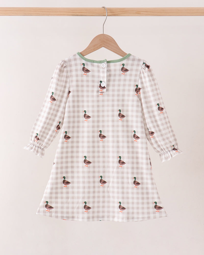 A long-sleeve baby dress with a beige and white gingham pattern and small duck prints, hanging on a wooden hanger against a plain light beige wall.