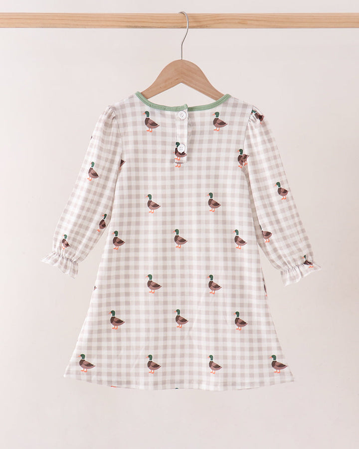 A long-sleeve baby dress with a beige and white gingham pattern and small duck prints, hanging on a wooden hanger against a plain light beige wall.