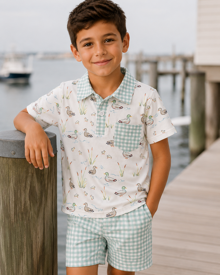 Child wearing a patterned shirt and shorts standing on a dock by water.