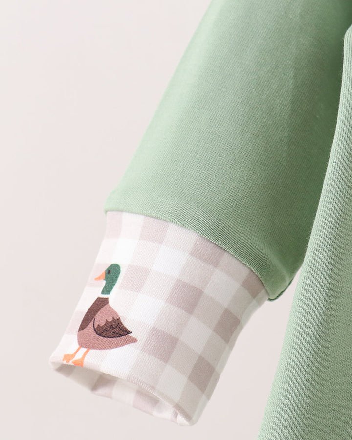 Close-up of a sage green long-sleeve top with a turned-up cuff showing a white and beige gingham pattern and a duck illustration.